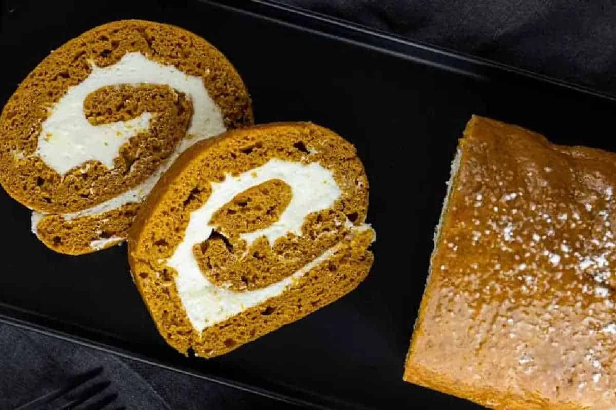 Two slices of pumpkin roll cake with cream cheese filling are displayed next to the remaining unsliced roll on a black rectangular plate.