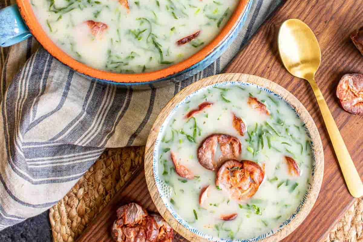A bowl and pot of creamy soup with slices of sausage and chopped greens, placed on a wooden surface with a gold spoon beside the bowl.