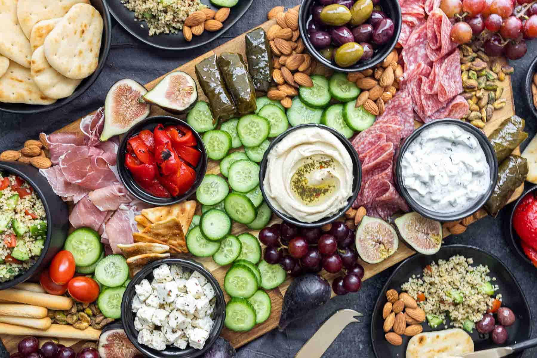 A platter with cucumber slices, hummus, feta cheese, olives, grapes, figs, pita bread, nuts, dolmas, meats, tzatziki, cherry tomatoes, and red peppers arranged on a wooden board.