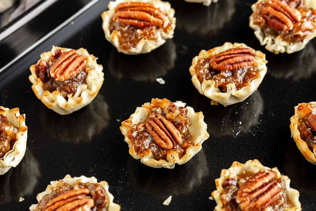 Mini pecan tarts with flaky crusts, filled with pecan pie filling and topped with pecan halves, arranged on a dark baking sheet.