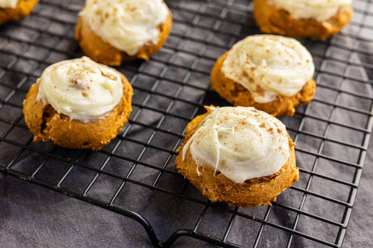 Pumpkin cookies with white icing and a dusting of spices are cooling on a black wire rack.