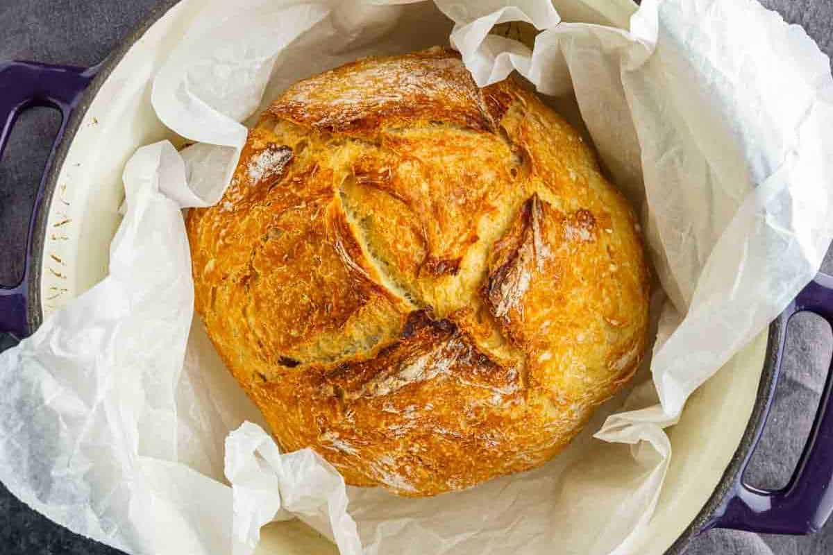 A round loaf of golden-brown artisan bread sits on parchment paper inside a Dutch oven.