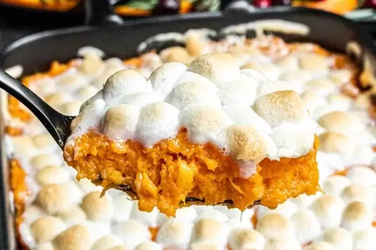 A serving spoon holds a portion of sweet potato casserole topped with toasted marshmallows above a baking dish filled with the same dish.