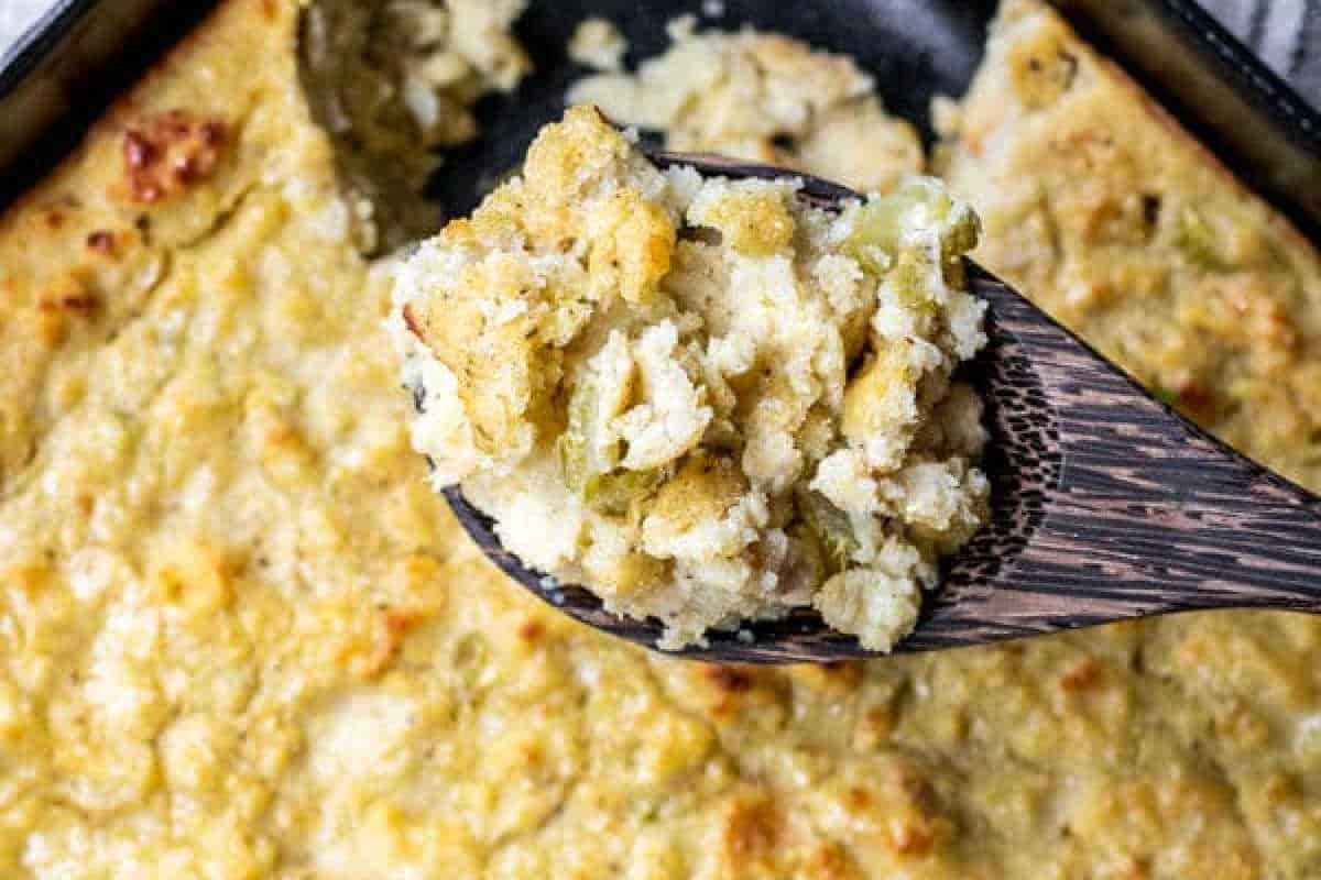 A close-up of a wooden spoon holding a serving of baked cornbread dressing above a casserole dish filled with the same dish.