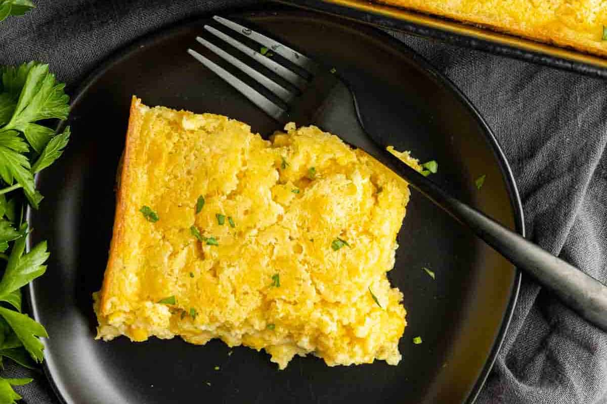 A slice of cheesy baked egg casserole on a black plate with a fork, garnished with chopped herbs, next to sprigs of fresh parsley.