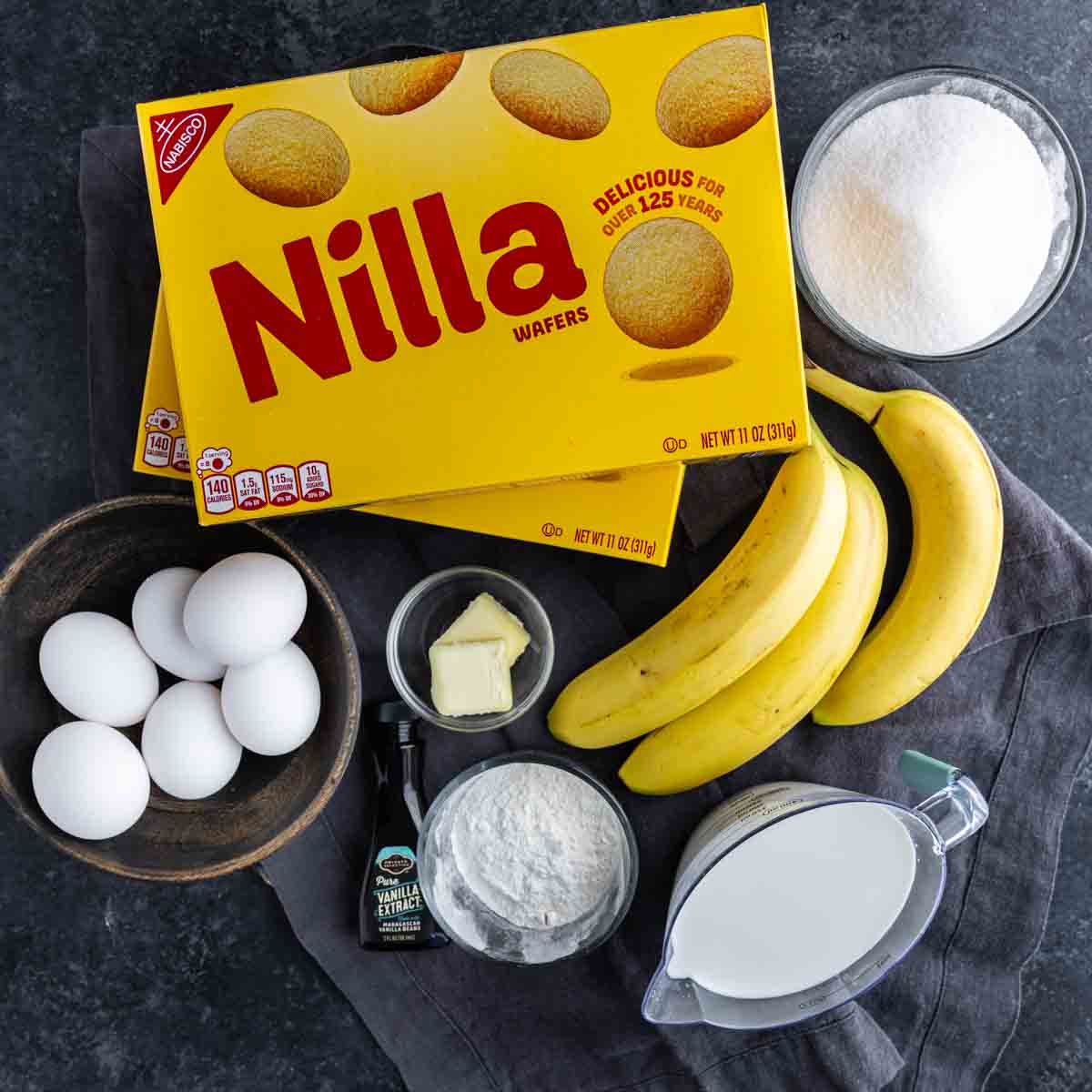 Box of Nilla Wafers, bananas, eggs, sugar, flour, milk, vanilla extract, and butter arranged on a dark surface with a gray towel—everything you need for an easy banana dessert or homemade vanilla pudding.