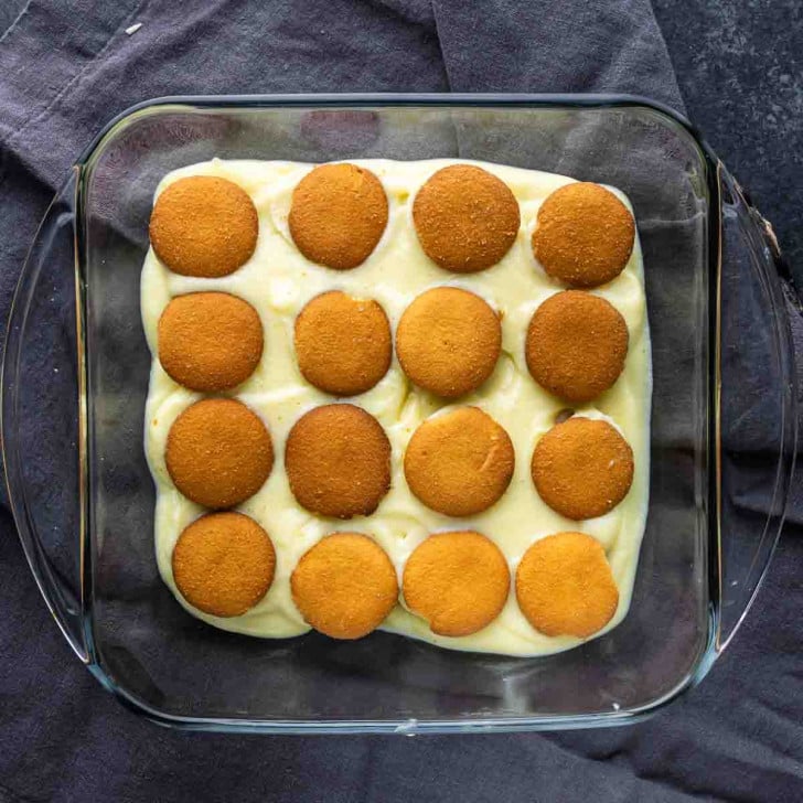 A glass baking dish filled with creamy homemade vanilla pudding is topped with 16 round vanilla wafer cookies, neatly arranged in a grid—an inviting and easy banana dessert.