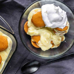 A glass bowl of classic banana pudding with vanilla wafers and a dollop of whipped cream sits next to a baking dish and a spoon on a dark cloth—an easy banana dessert perfect for any occasion.