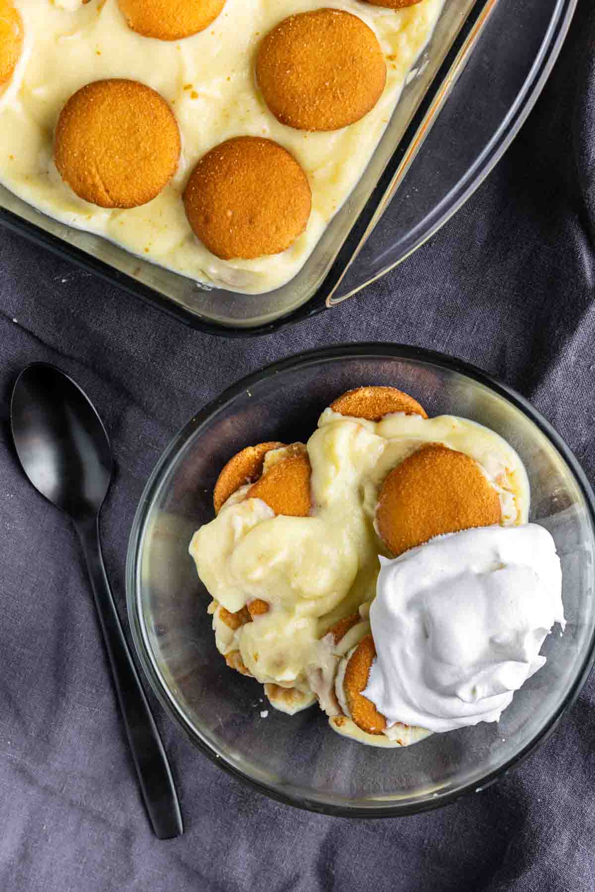 A bowl of classic banana pudding topped with vanilla wafers and whipped cream sits beside a baking dish of this easy banana dessert, with a black spoon resting on a gray cloth.