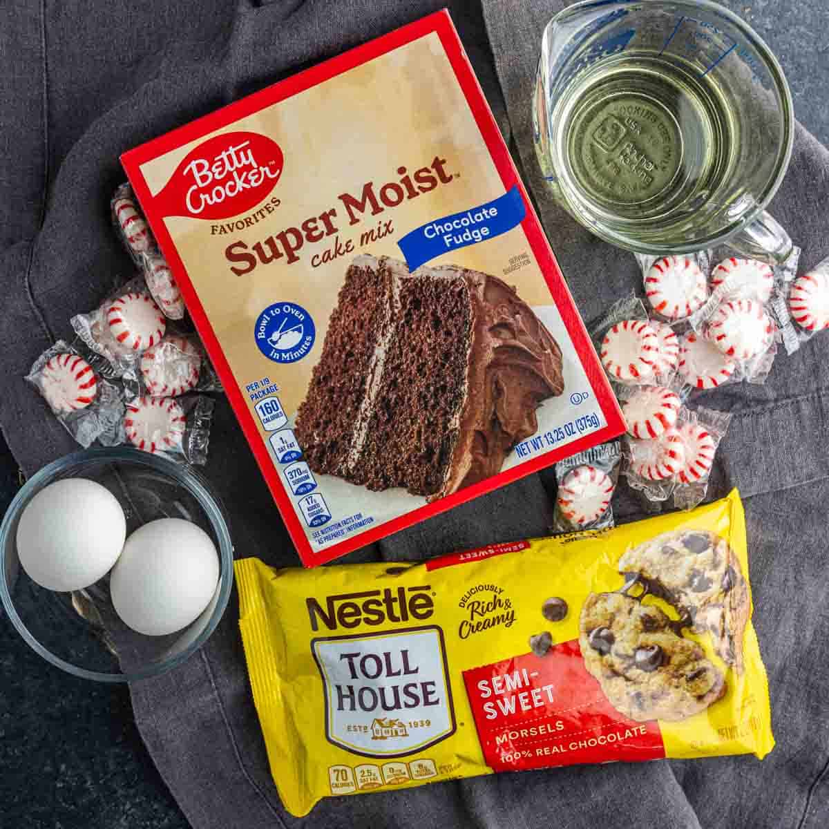 A box of cake mix, semi-sweet chocolate chips, eggs, oil, and wrapped peppermint candies are arranged for easy peppermint cake mix cookies on the counter.