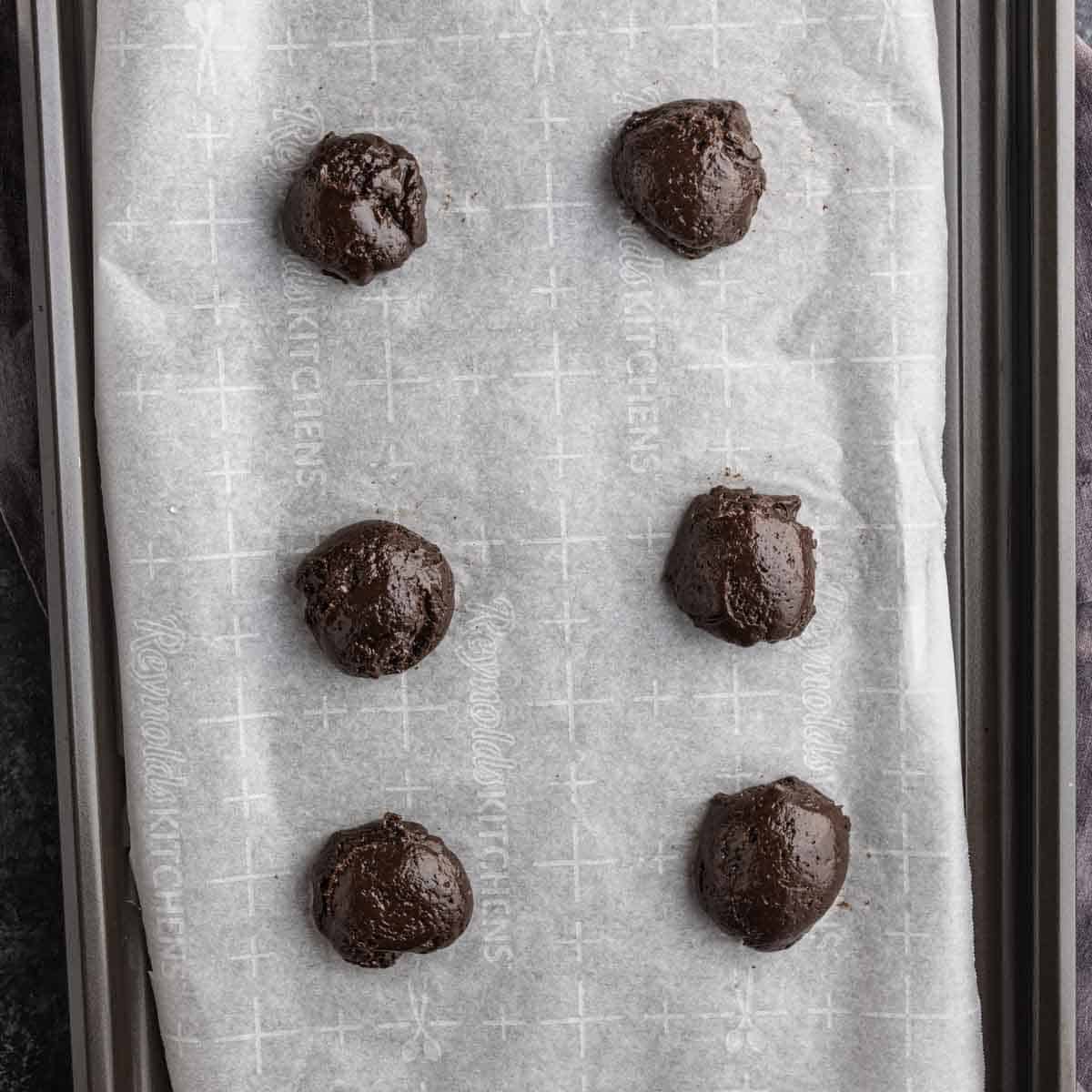Six scoops of chocolate peppermint cake mix cookies dough are placed on a parchment-lined baking sheet, spaced evenly apart in two columns.