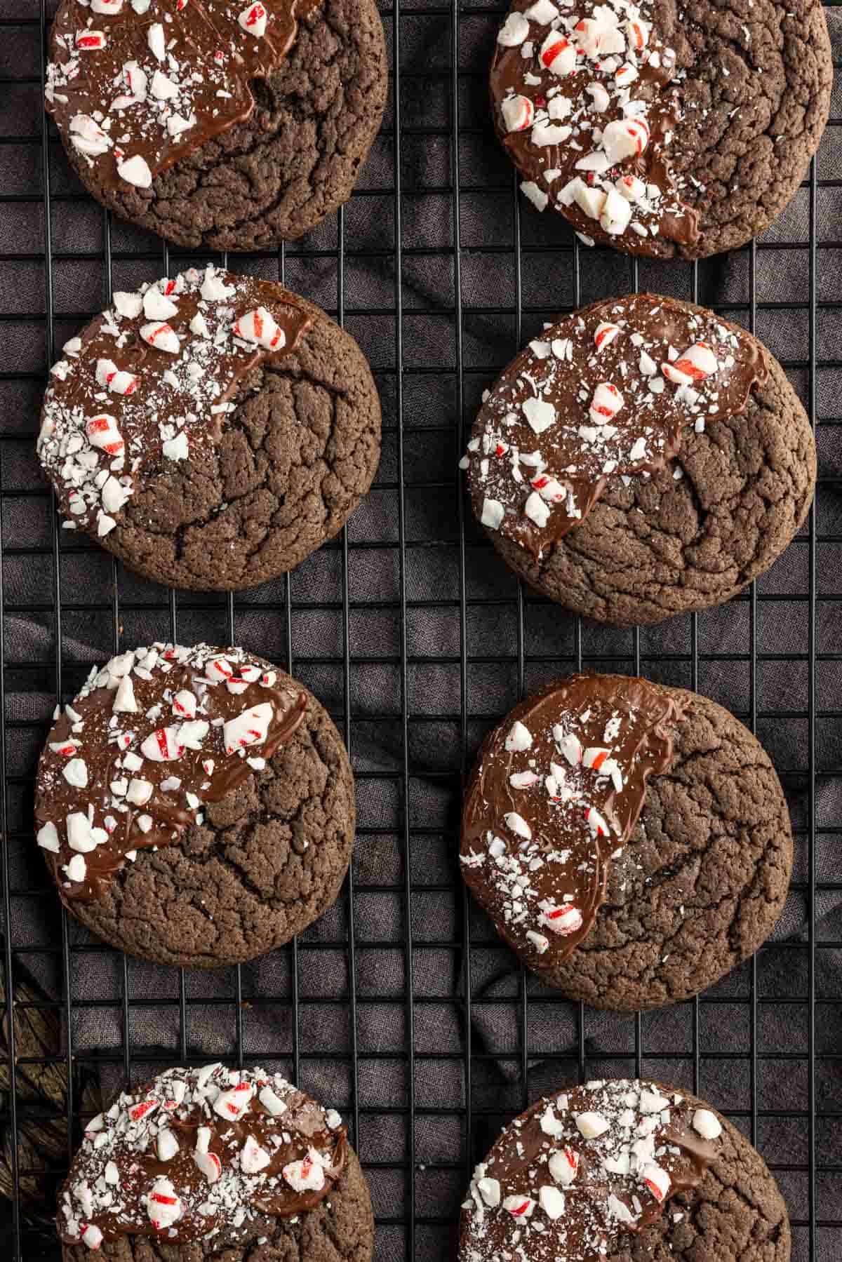 Eight chocolate cookies, some dipped in chocolate and sprinkled with crushed peppermint, are cooling on a black wire rack—these easy peppermint cake mix cookies are perfect for the holidays.