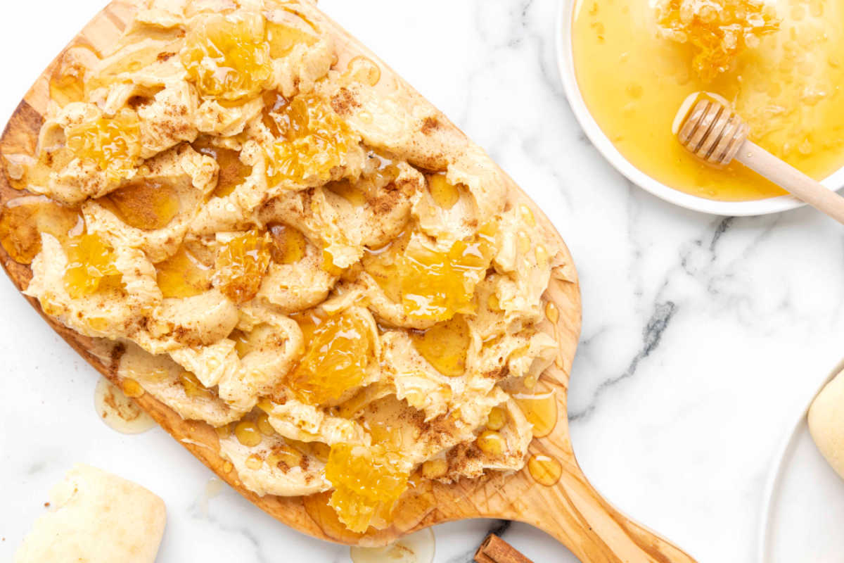 Torn pieces of bread drizzled with honey on a wooden serving board, next to a bowl of honey with a honey dipper on a marble surface.