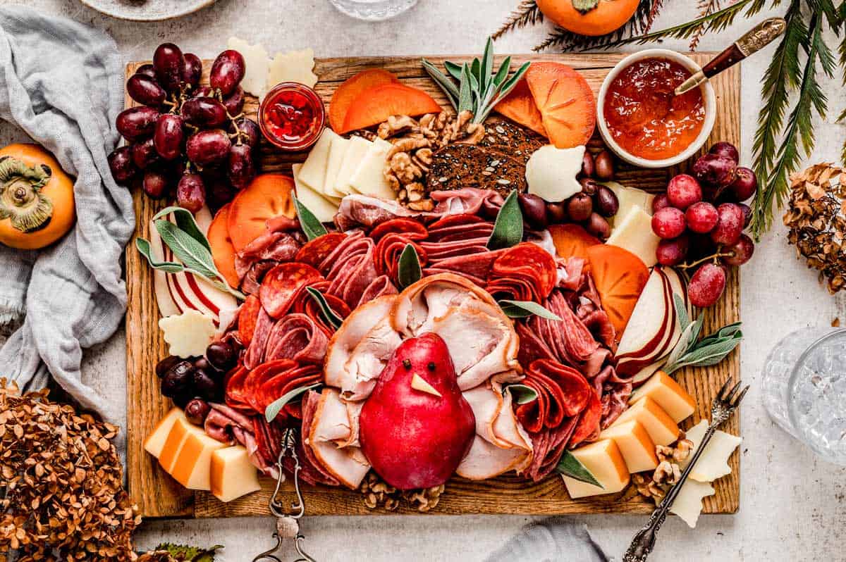 A wooden board with assorted sliced meats, cheeses, grapes, persimmon slices, walnuts, crackers, red jam, and a whole pomegranate in the center, arranged decoratively.