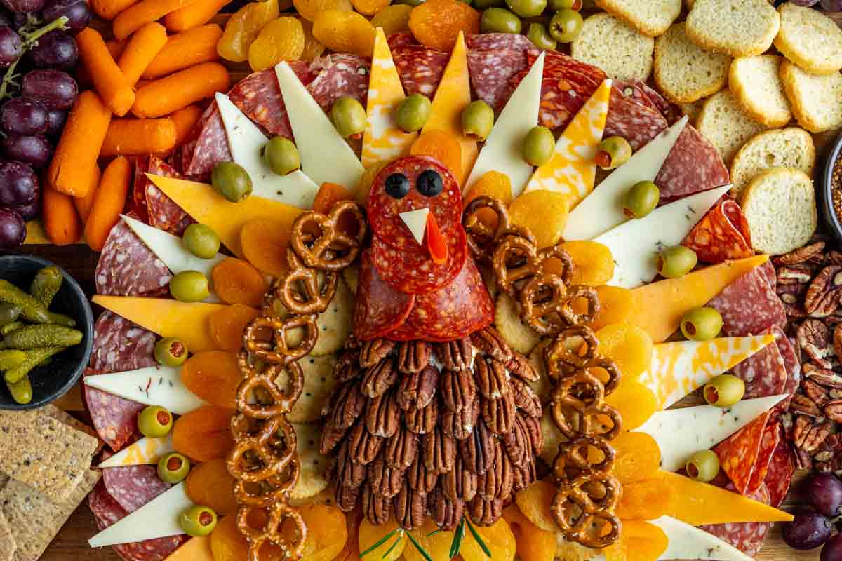 A charcuterie board arranged to resemble a turkey, with meats, cheeses, olives, pretzels, pecans, crackers, grapes, carrots, and dried apricots.