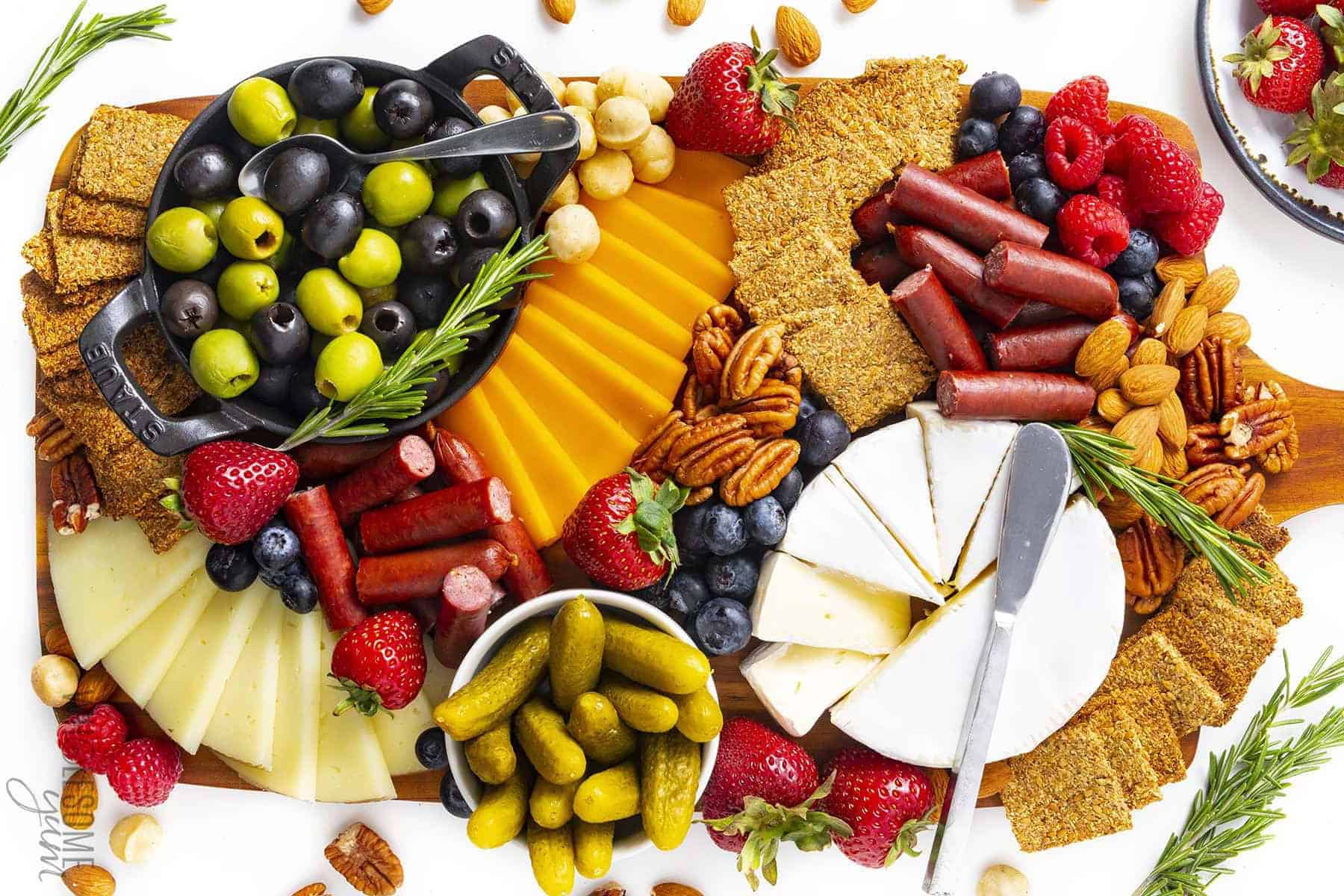 A charcuterie board with assorted cheeses, crackers, cured meats, olives, strawberries, blueberries, pickles, and mixed nuts, garnished with rosemary sprigs.