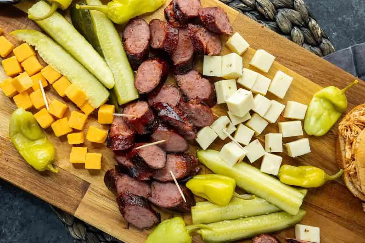A wooden board with sliced sausage, pickle spears, pepperoncini, and cubes of cheddar and white cheese, arranged neatly.
