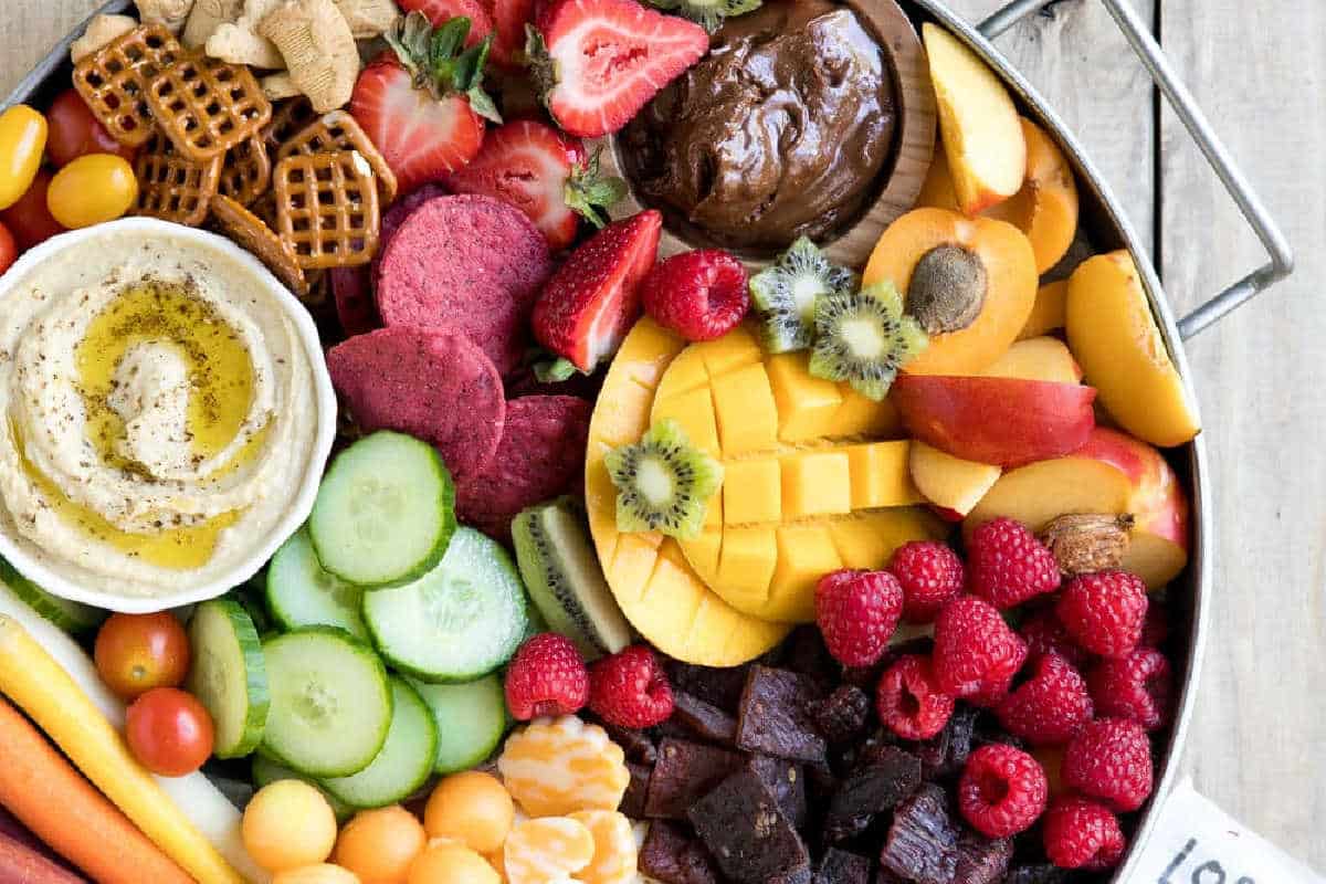 A platter with assorted fresh fruits, sliced vegetables, pretzels, crackers, cheese, hummus with olive oil, and a chocolate dip.