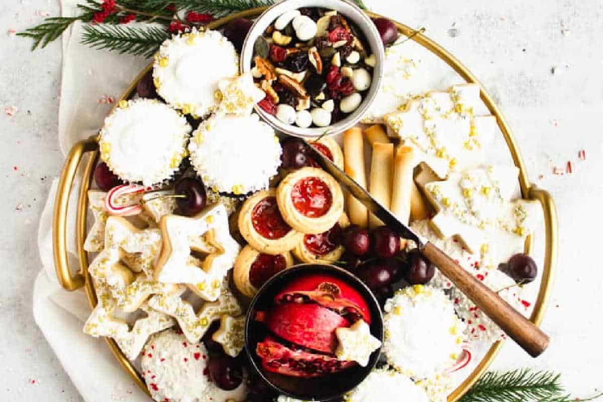 A gold tray holds assorted Christmas cookies, jam tarts, chocolate, nuts, grapes, a pomegranate half, and festive decorations, with a knife resting on the tray.