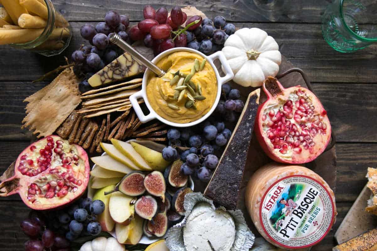 A charcuterie board with sliced fruits, grapes, figs, crackers, dip, cheese, pomegranate halves, a small white pumpkin, and a container of Petit Basque cheese on a wooden table.