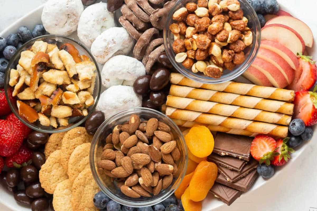 A platter with assorted snacks including almonds, cookies, chocolate, rolled wafers, dried apricots, apples, strawberries, blueberries, sugared nuts, and powdered donuts.