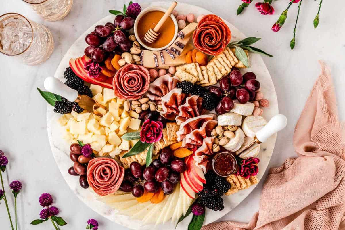 A round charcuterie board with assorted cheeses, meats, grapes, blackberries, crackers, nuts, honey, jam, and garnished with edible flowers.
