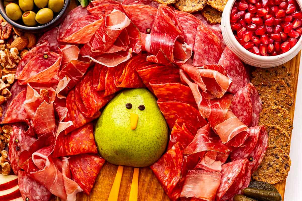 A charcuterie board arranged to resemble a turkey, with a pear as the body, slices of cured meats as feathers, and sides like olives, pomegranate seeds, nuts, and crackers.