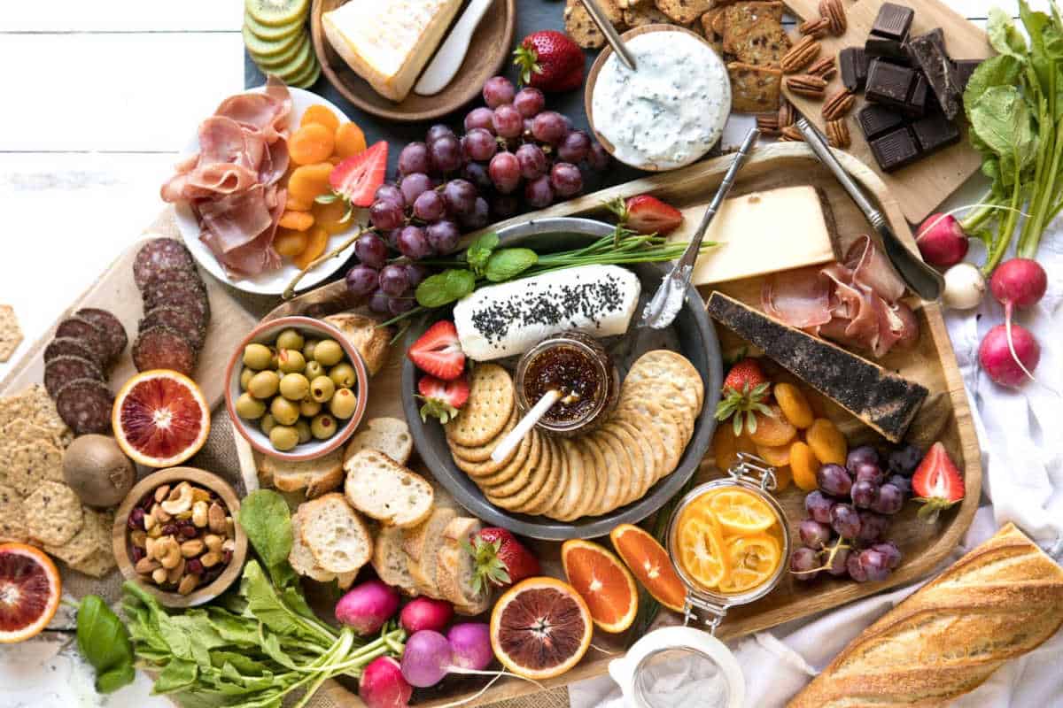 A charcuterie board with assorted cheeses, crackers, meats, grapes, olives, nuts, fresh vegetables, fruit slices, dip, and chocolate, arranged on a wooden surface.
