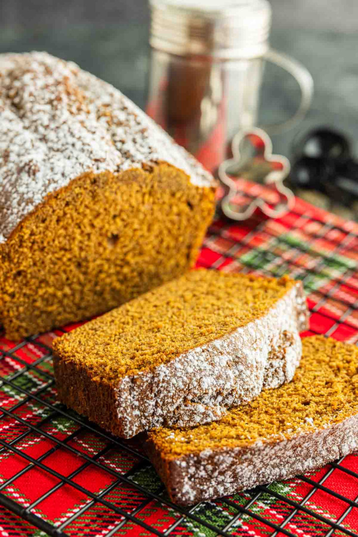 A loaf of pumpkin bread with two slices cut, dusted with powdered sugar, sits on a red plaid cloth with a sifter and cookie cutter in the background.