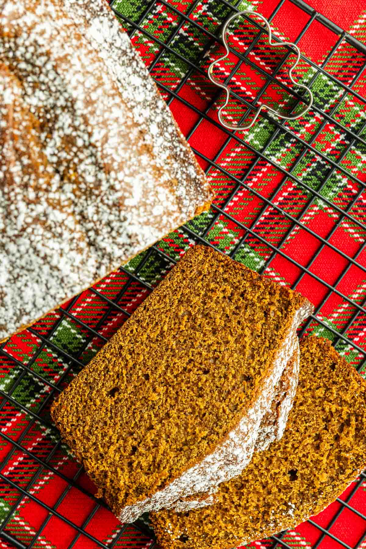 Sliced homemade gingerbread loaf dusted with powdered sugar rests on a red and green plaid cloth, with a metal cookie cutter nearby.