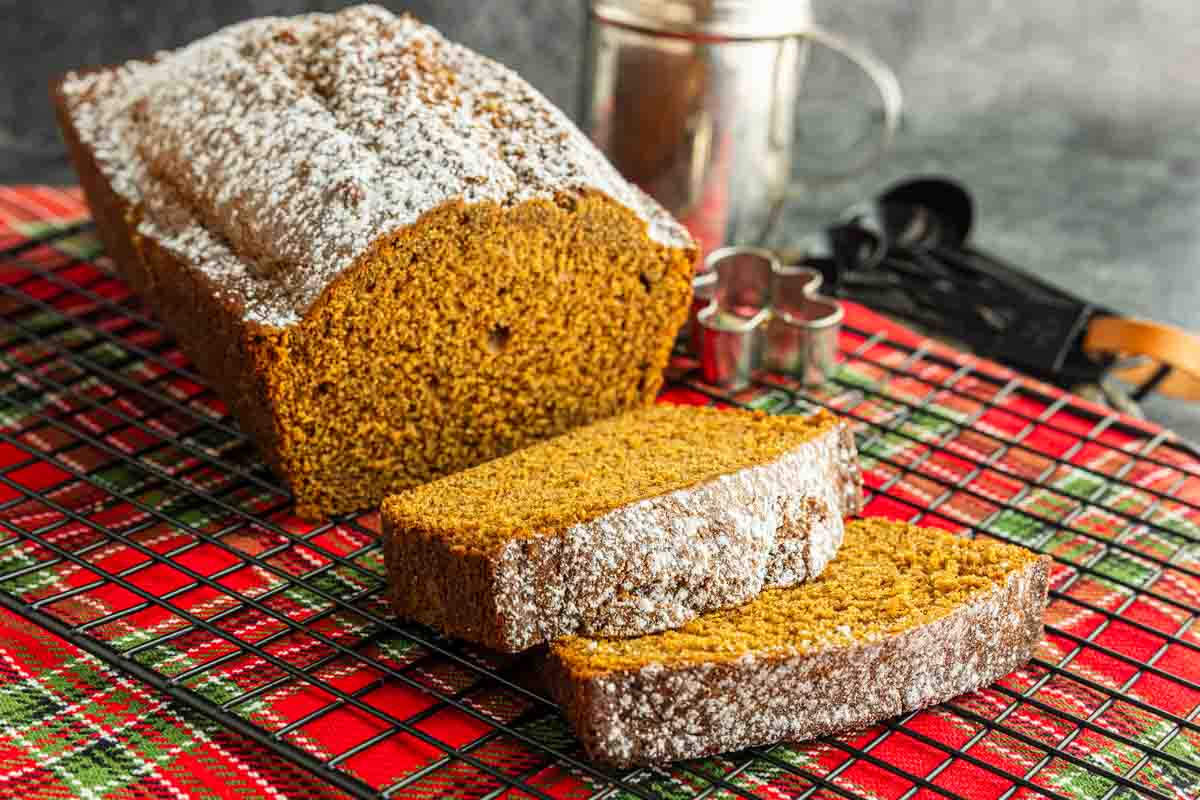 A homemade gingerbread loaf dusted with powdered sugar, with two slices cut, sits on a wire rack over a red plaid cloth. Baking utensils are in the background.