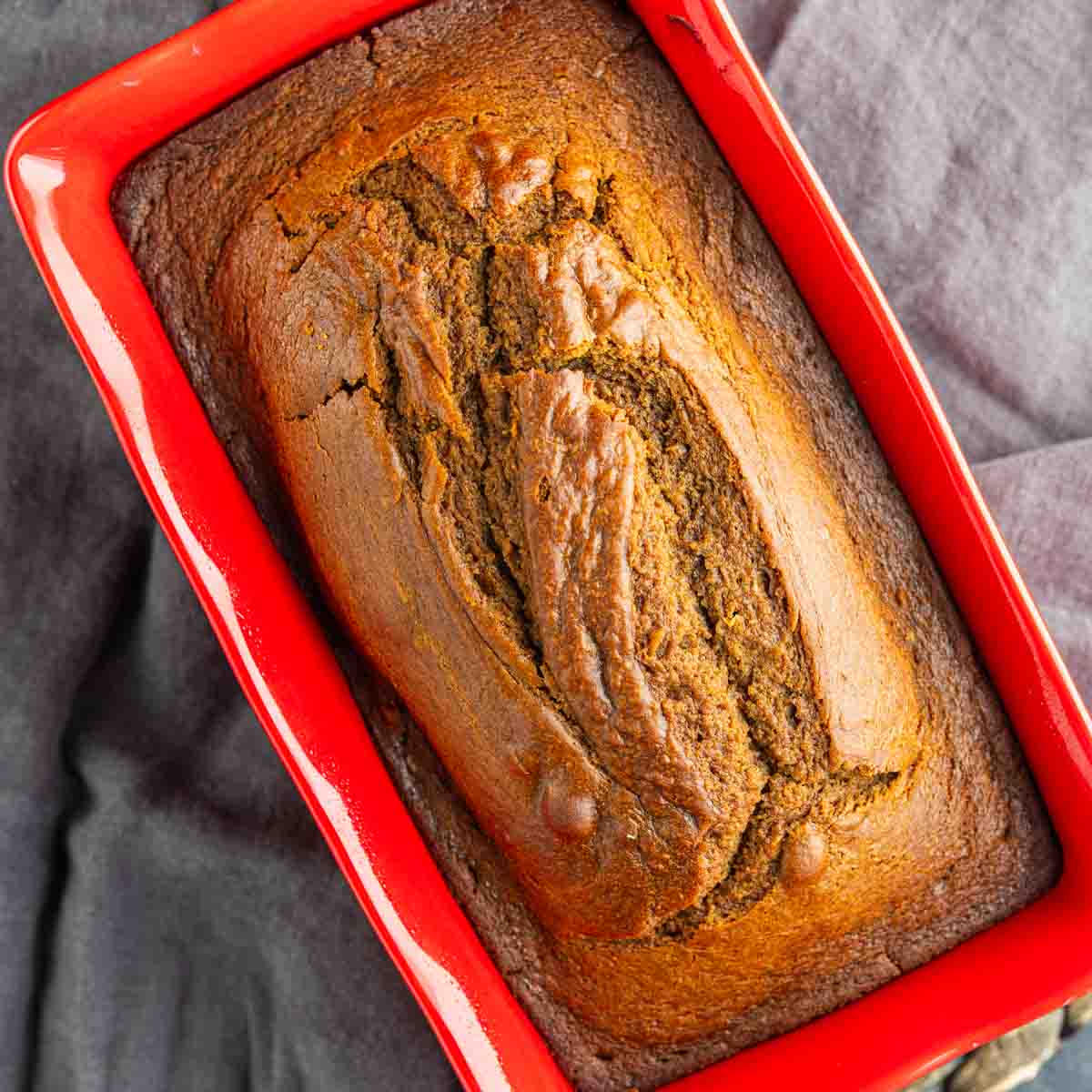 A loaf of baked gingerbread pound cake in a red rectangular pan, resting on a dark cloth background.