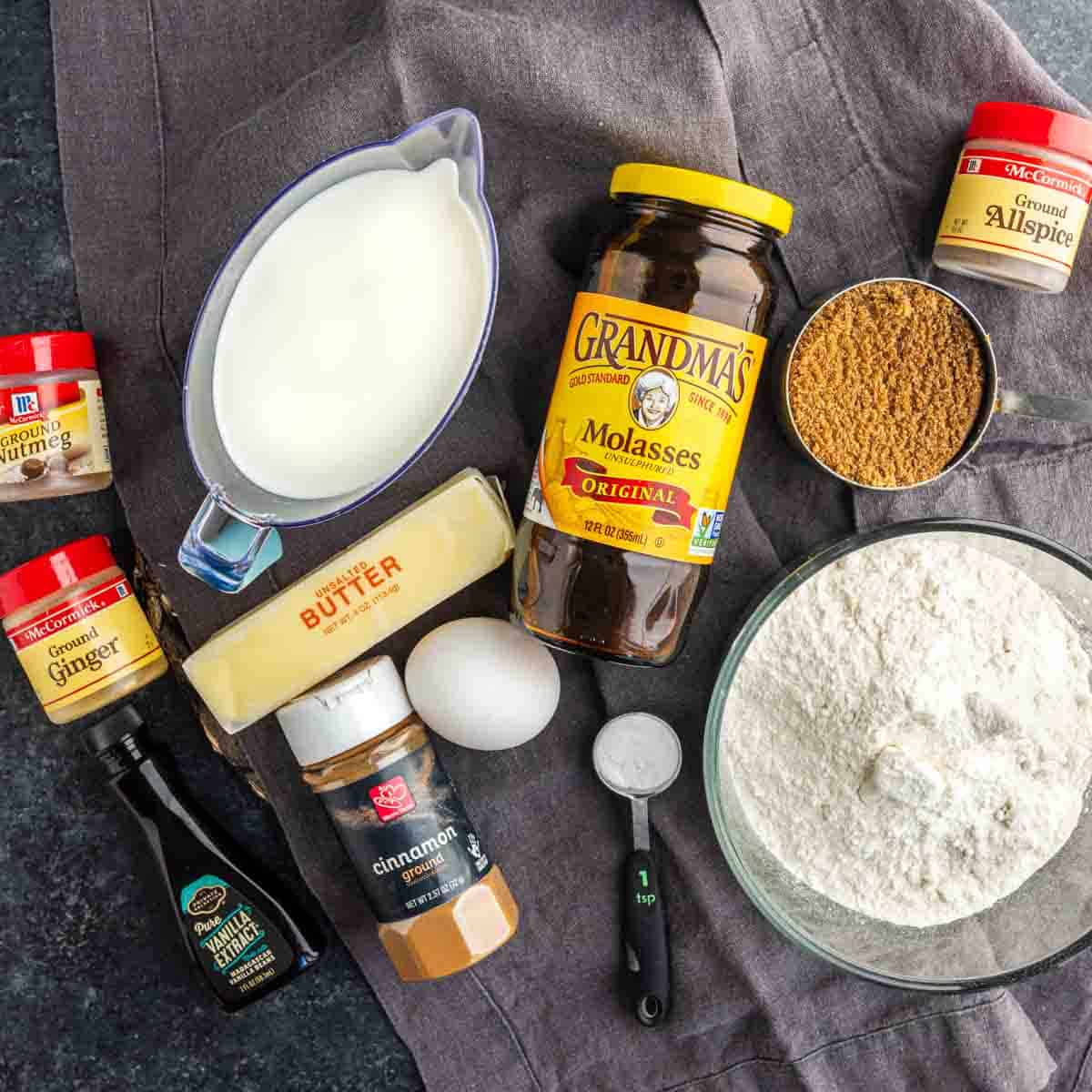 Baking ingredients on a gray cloth, including flour, brown sugar, butter, spices, molasses, milk, an egg, and vanilla extract—perfect for making a moist gingerbread cake or a homemade gingerbread loaf inspired by Starbucks.