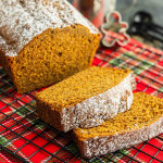 A loaf of pumpkin bread with two slices cut, topped with powdered sugar, sits on a red plaid cloth.