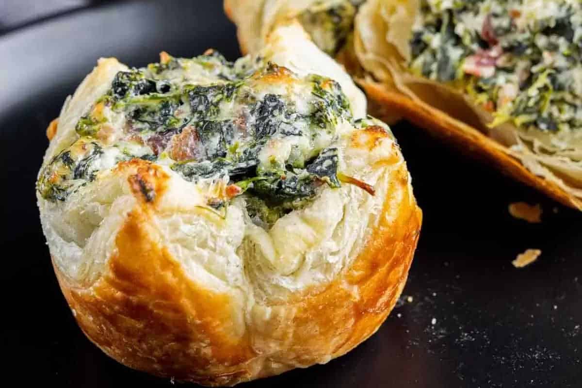 A close-up of a baked pastry filled with creamy spinach and cheese, with a second pastry cut open to reveal the filling—perfect as holiday appetizers or for your New Year’s Eve party food spread.