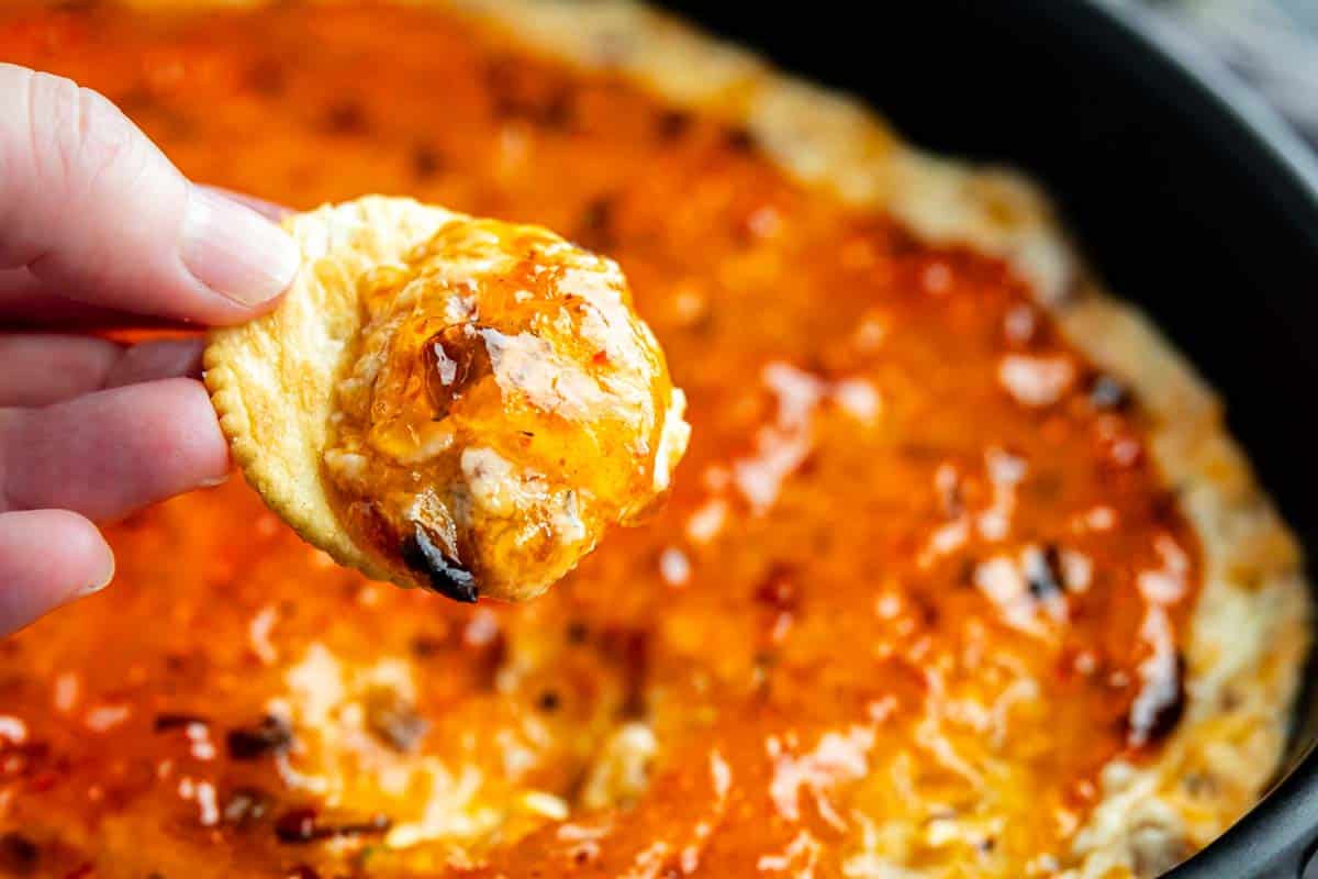 Hand holding a cracker with a serving of hot pepper jelly dip in front of a pan filled with the same savory appetizer.