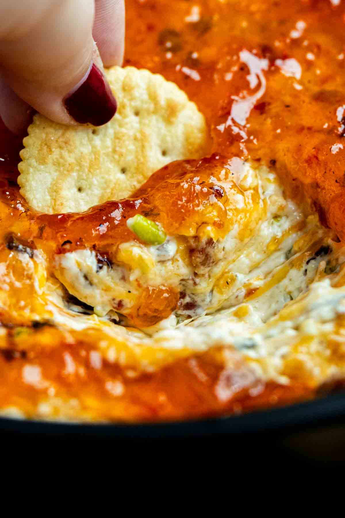 A hand dips a cracker into a hot, cheesy, baked cheese dip recipe with a bubbly golden topping—perfect for serving as a crowd-pleasing hot pepper jelly appetizer.