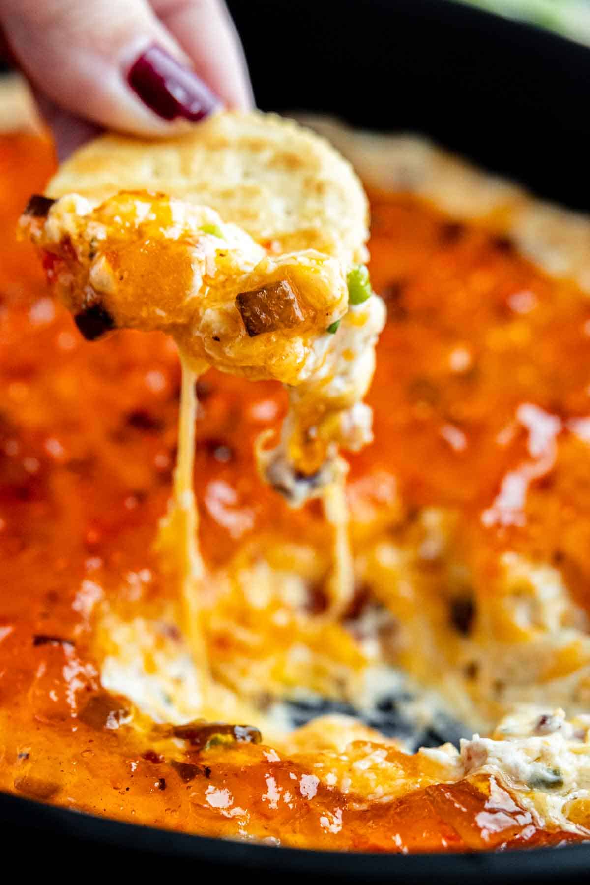 A hand holds a tortilla chip dipped into a cheesy, baked bacon and cream cheese dip, with melted cheese stretching from the chip to the dish.