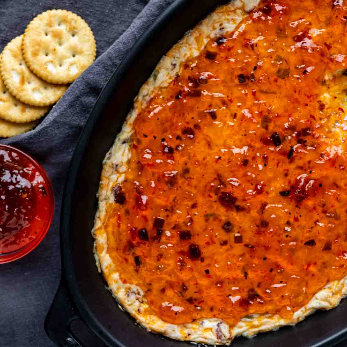 Baked cheese dip topped with hot pepper jelly in a black baking dish, served with round crackers and a small bowl of extra jelly on the side—a perfect hot pepper jelly appetizer for any gathering.