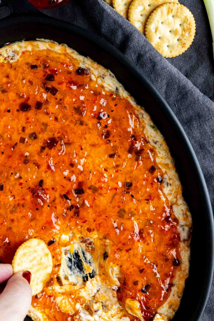 A hand dipping a round cracker into a creamy cheese dip topped with orange sweet chili sauce in a black dish.