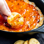 A hand dips a round cracker into a baking dish filled with a glossy, orange-colored spread or dip.