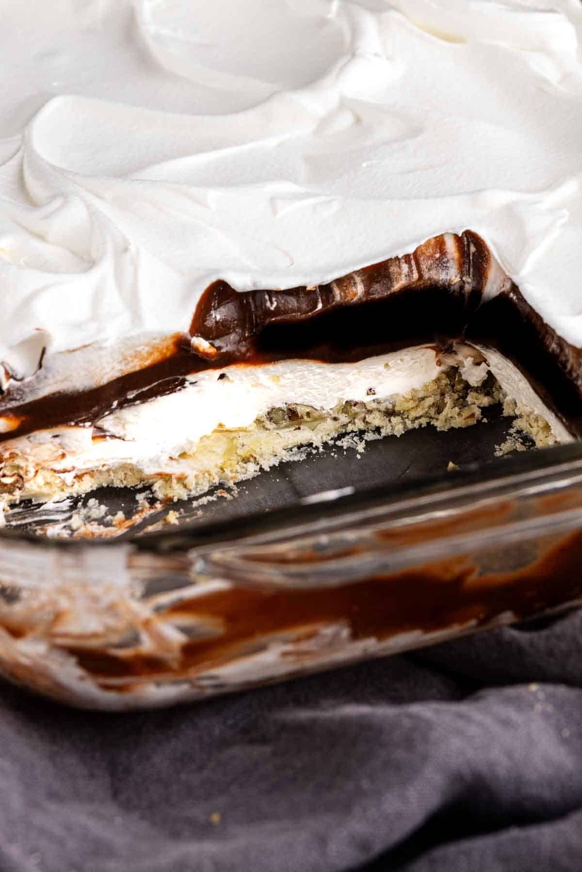 A glass baking dish with a layered chocolate lasagna dessert featuring a whipped topping, rich chocolate layer, and crumbly crust, with a section missing to show the delicious layers.