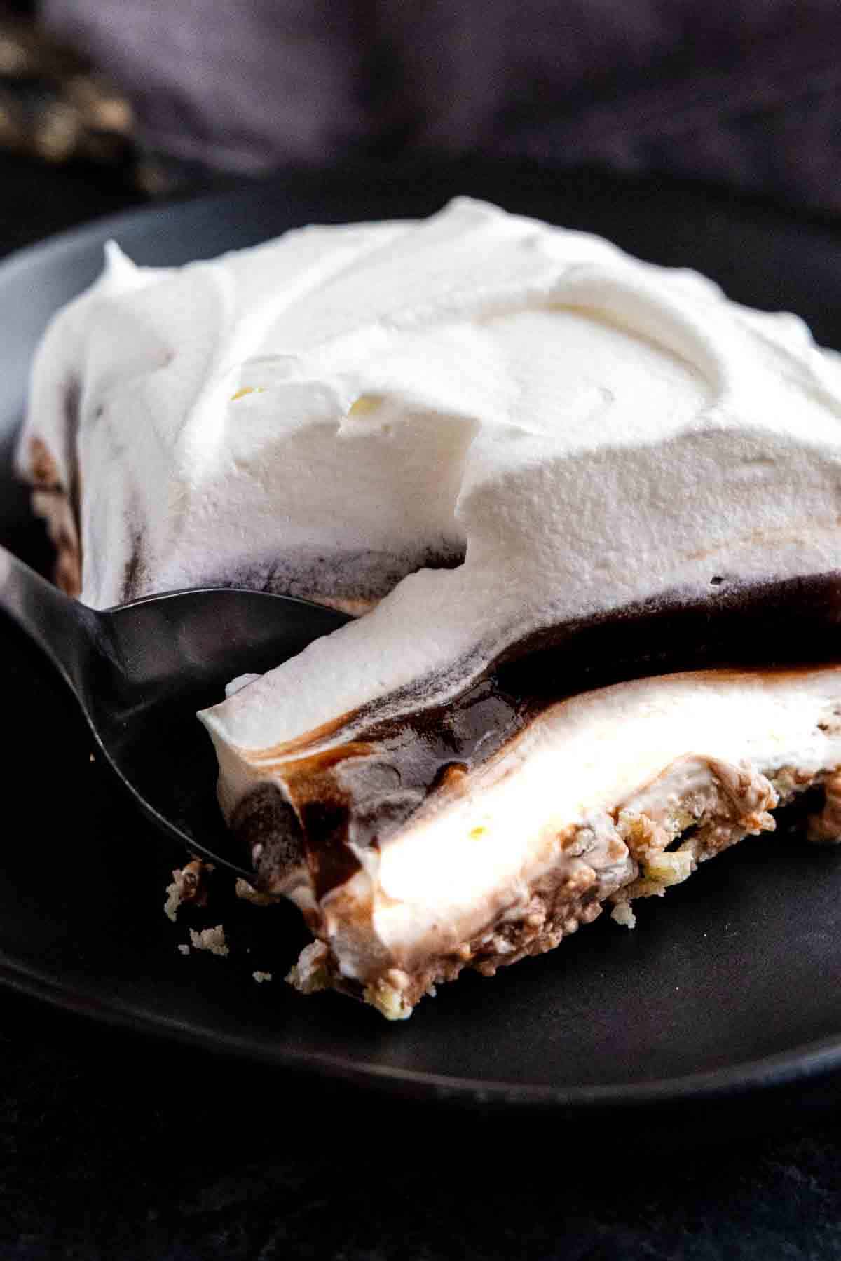 A close-up of a layered chocolate pudding dessert with whipped cream, rich chocolate, and a crumbly crust on a black plate, as a spoon takes a bite.