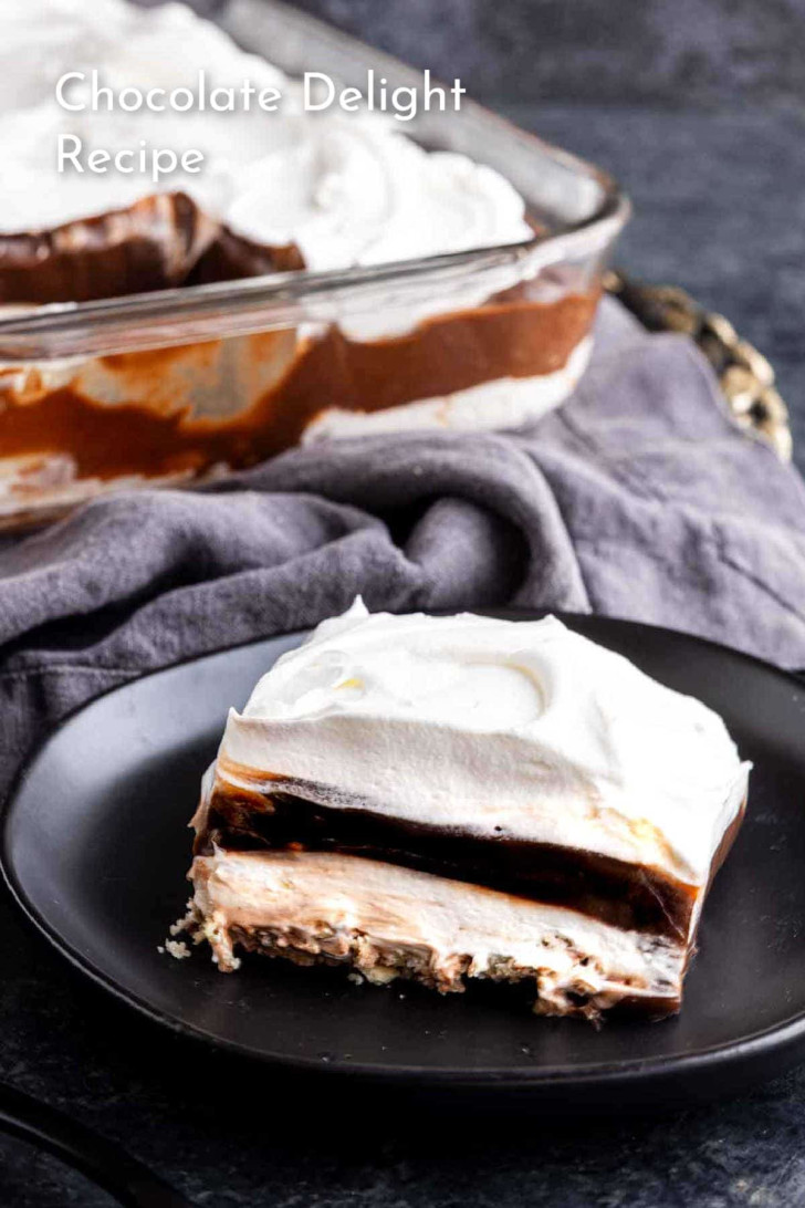 A slice of layered chocolate delight dessert with whipped topping on a black plate, with the rest of the dessert in a glass dish in the background.