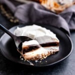 A fork holds a bite of layered chocolate dessert with whipped topping, resting on a black plate.