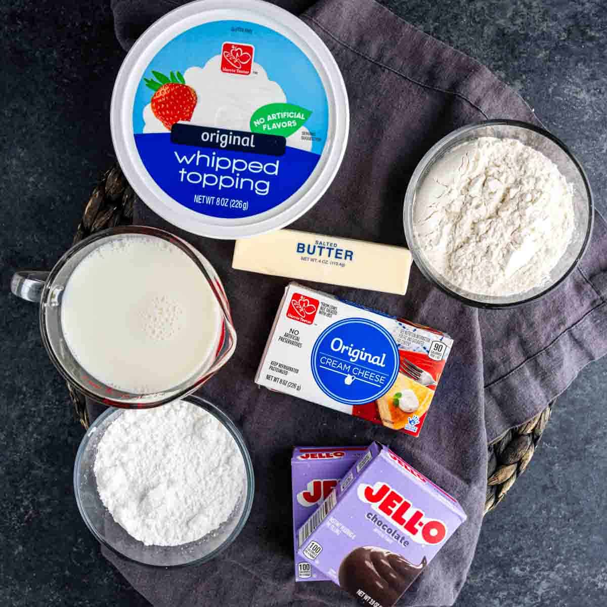 Ingredients including whipped topping, flour, stick of butter, milk, cream cheese, powdered sugar, and chocolate pudding mix arranged on a dark surface—perfect for making a rich layered chocolate pudding dessert like Chocolate Delight.