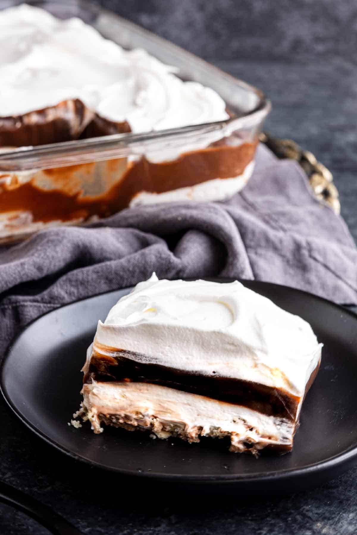 A slice of luscious chocolate lasagna dessert with whipped cream, rich chocolate, and cream filling sits on a black plate, while the rest of the layered treat awaits in a glass dish in the background.