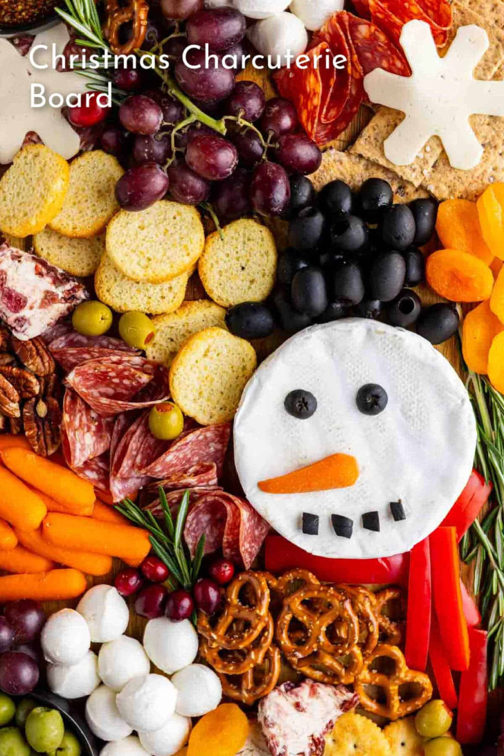 A festive charcuterie board with meats, cheese, crackers, grapes, carrots, olives, pretzels, dried fruit, and a snowman made of cheese and vegetables. Text reads "Christmas Charcuterie Board.