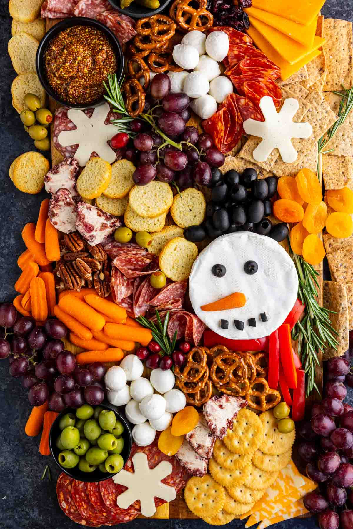 A festive charcuterie board with assorted crackers, cheeses, meats, fruits, veggies, and a snowman-shaped cheese decorated with olives, carrots, and peppers makes the perfect holiday charcuterie board for your Christmas gatherings.
