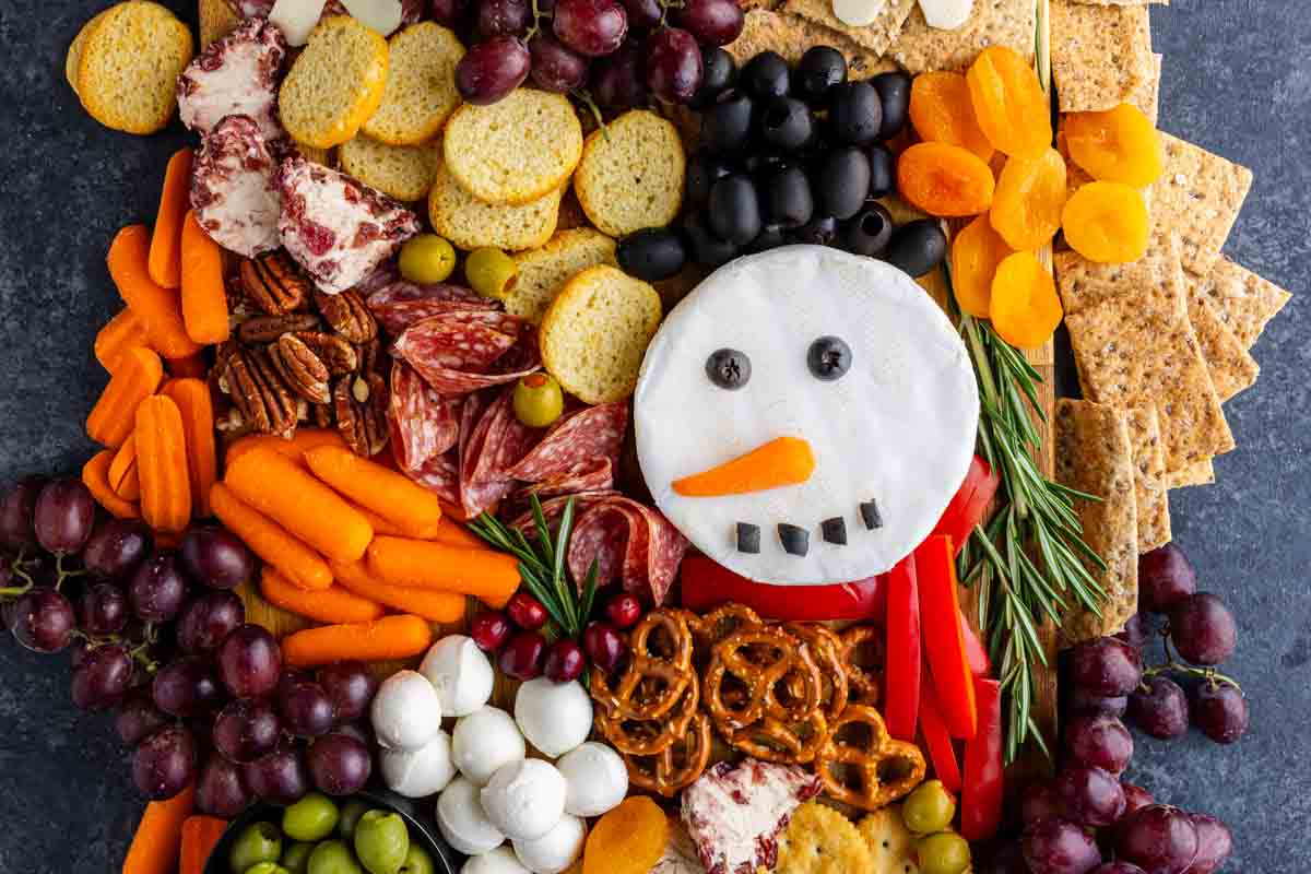 A festive charcuterie board with meats, cheeses, crackers, fruits, nuts, and veggies features a snowman face made from round cheese topped with olives and a carrot—perfect for your Christmas charcuterie board.